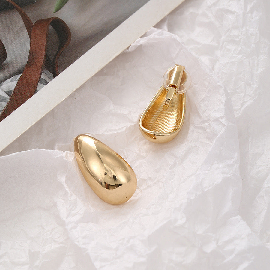 Gold tone chunky drop clip-on earrings with a minimalist bean shape, crafted from a copper alloy and featuring a secure hinge-back closure, showcased on a crinkled white paper background for an elegant presentation.