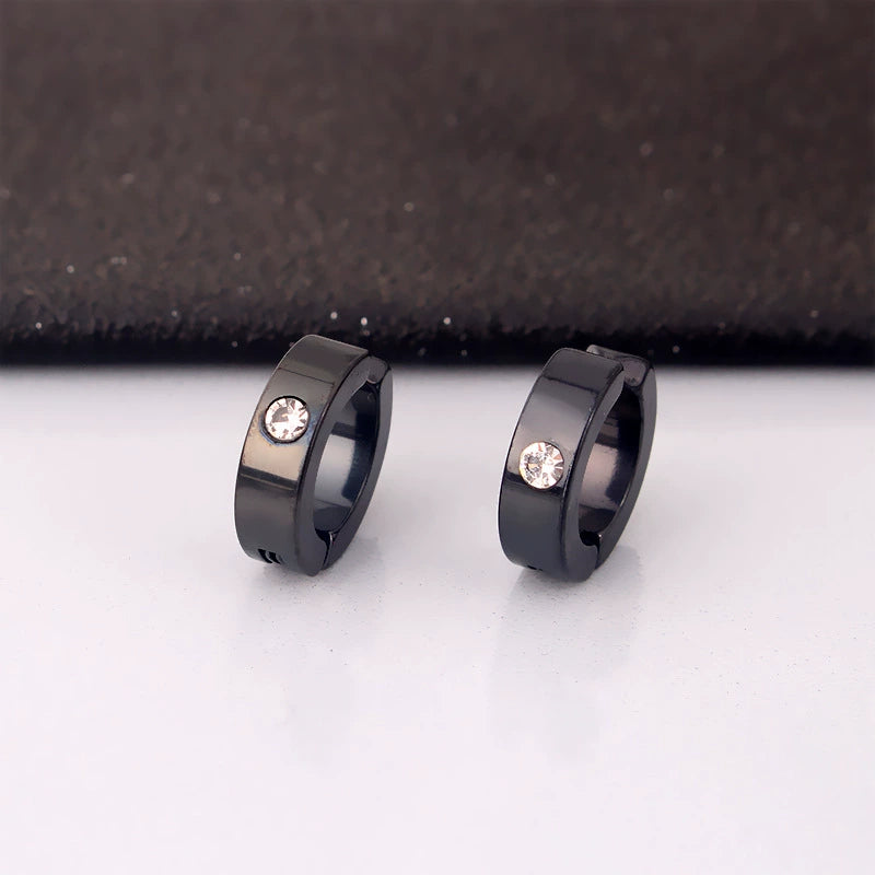 A pair of black minimalist clip-on huggie hoops with a single zircon stone, displayed on a white surface against a dark background.