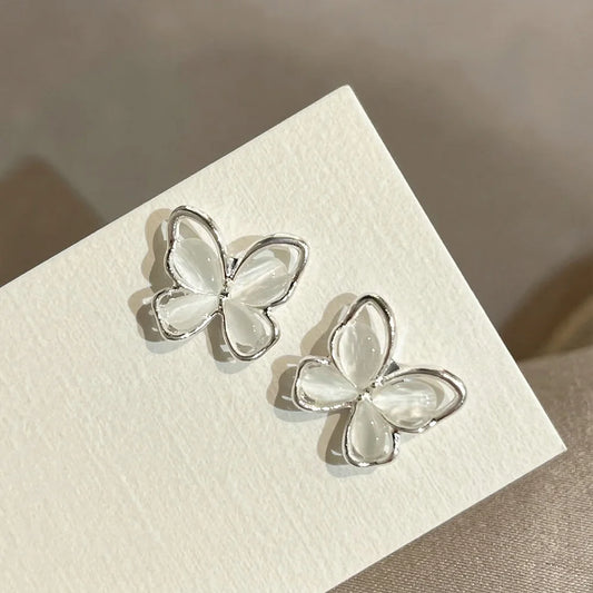 Two iridescent silver butterfly stud earrings positioned on a textured white card to highlight the light-catching quality of the wings.