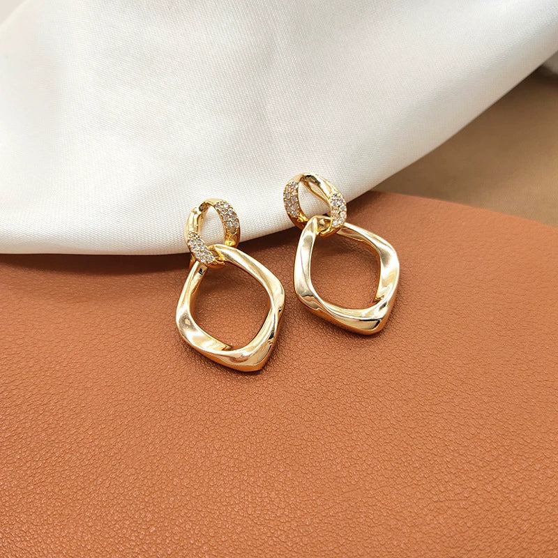Gold geometric thombus clip-on earrings on a brown surface with a white cushion in the background