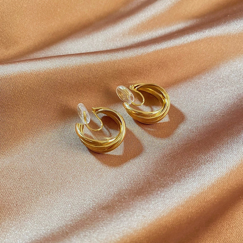 A pair of layered wave gold clip-on hoop earrings with an invisible resin clip, displayed on a shimmering satin background