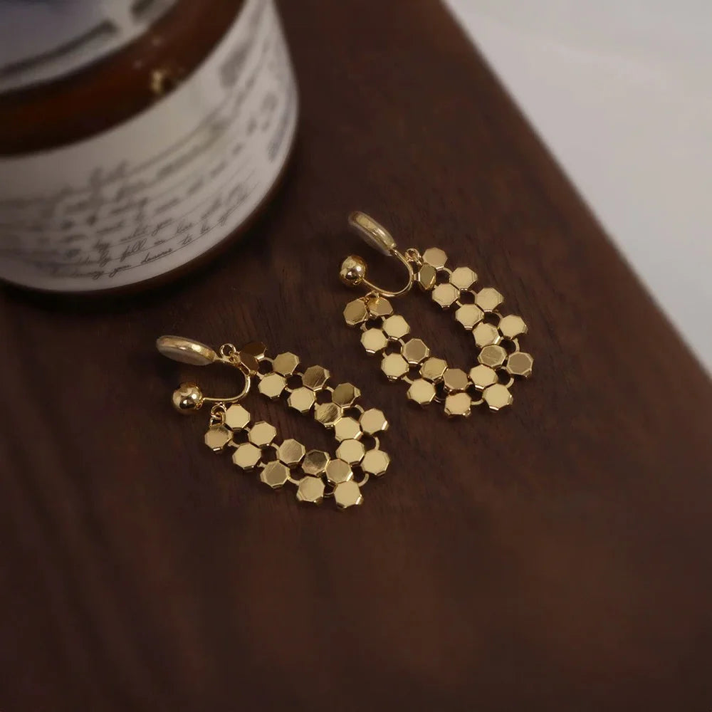 Geometric gold hexagonal clip-on earrings displayed on a rich, dark wood surface.