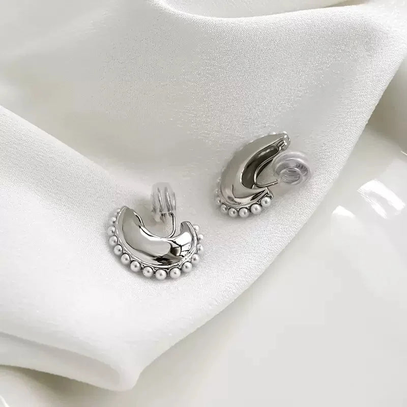 Angled view of the silver Luna pearl crescent clip-on earrings on a white satin cloth, showing the detail of the clip-on mechanism with silicone pads.