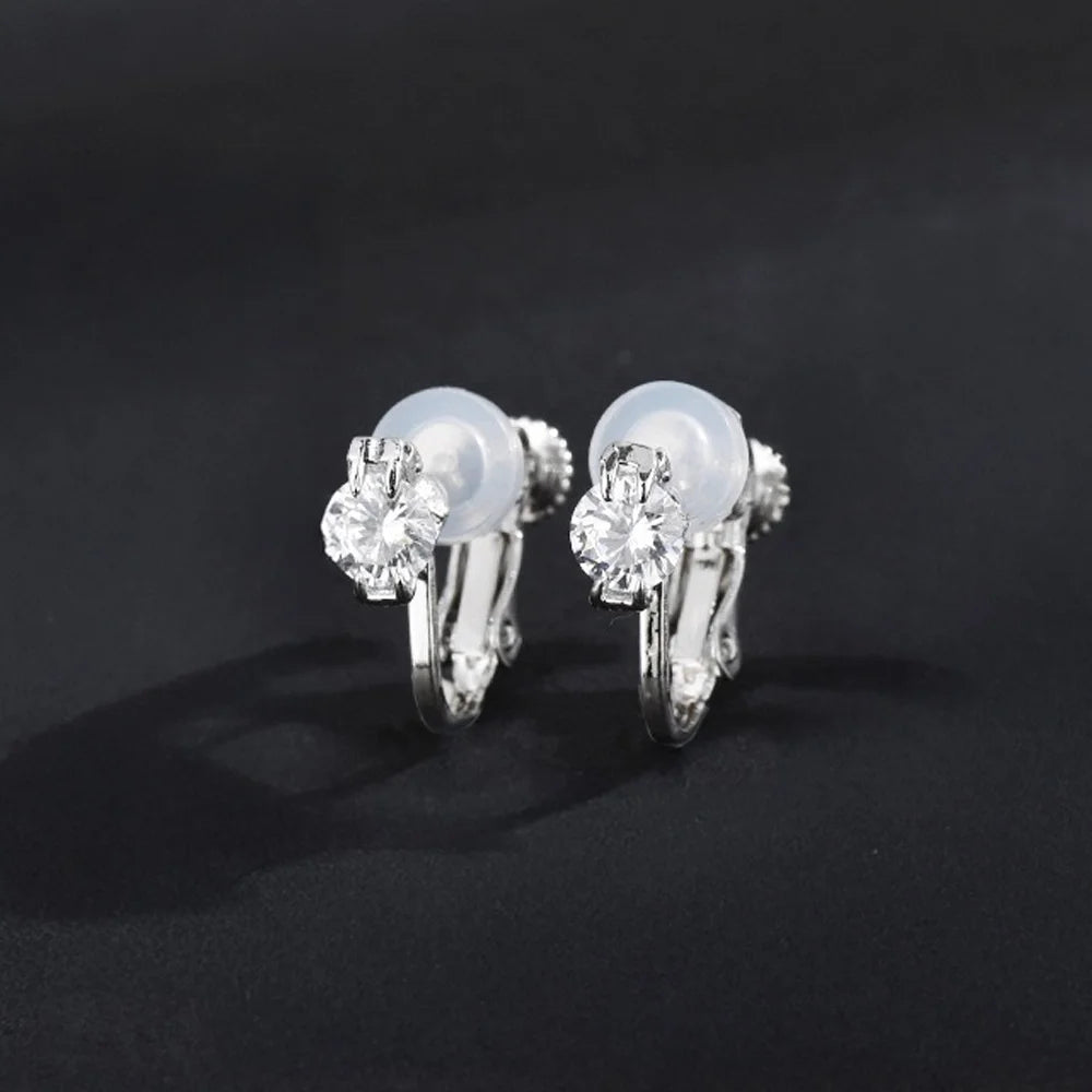 A pair of silver-tone solitaire clip-on stud earrings with clear cubic zirconia stones and cushioned clips, displayed on a black background.