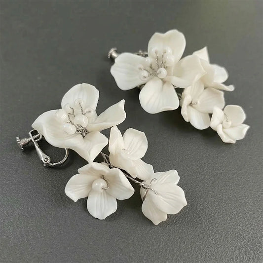 Close-up top-down view of the pair of detailed handcrafted white porcelain-style flower cascading clip-on bridal earrings resting on a dark grey textured surface, highlighting the pearl embellishments and silver screw-back closures.