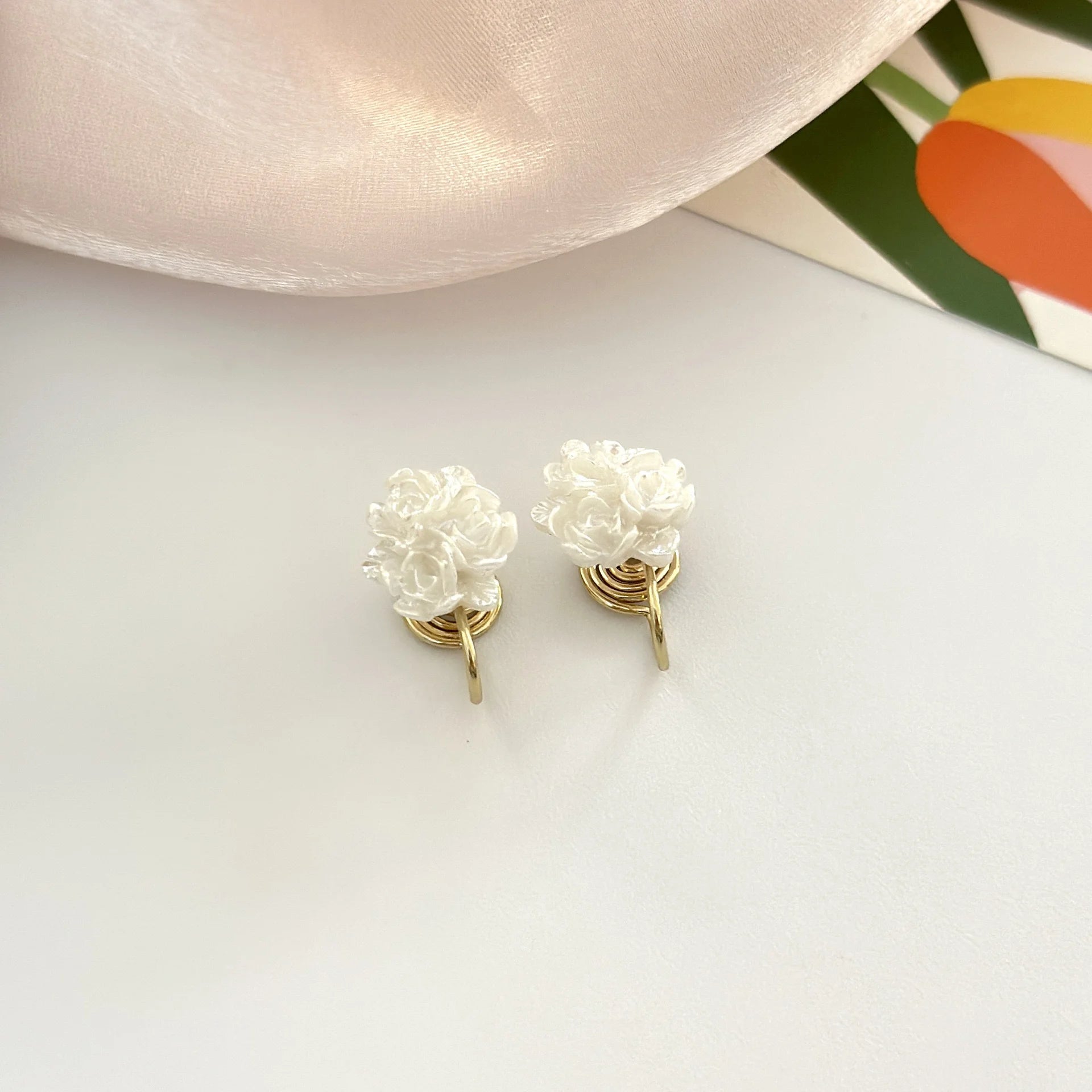 A pair of white resin flower clip-on earrings with comfortable mosquito coil clips displayed on a white surface next to a satin cloth.
