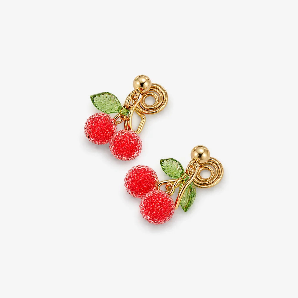 A pair of gold-plated cherry drop earrings with textured red cherries, green leaves, and a clip on back closure, shown on a light gray background.