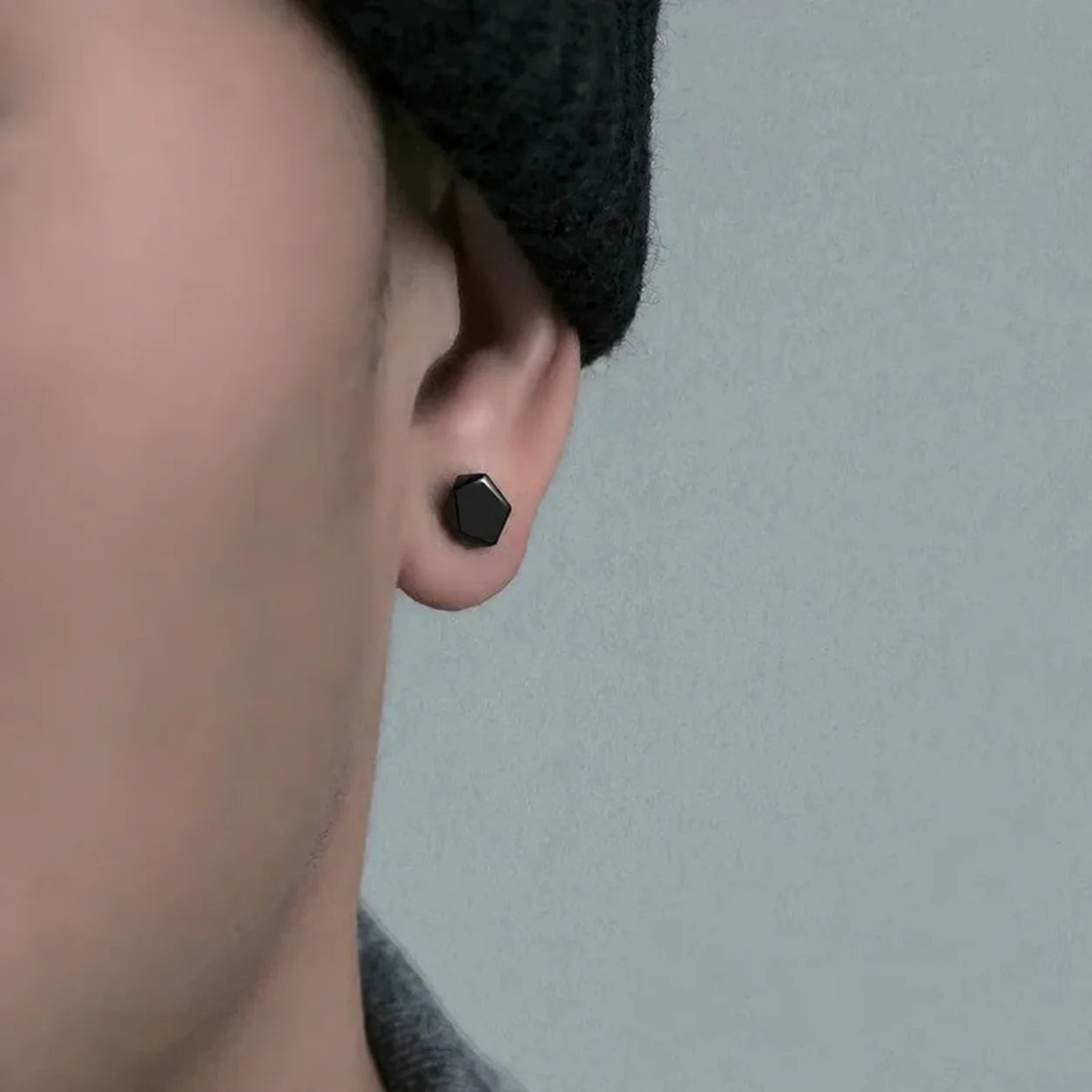A close-up of a person's ear wearing a small matte black hexagonal stud earring, with part of a dark knit beanie and gray shirt visible against a soft light gray background.