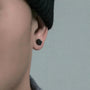 A close-up of a person's ear wearing a small matte black hexagonal stud earring, with part of a dark knit beanie and gray shirt visible against a soft light gray background.