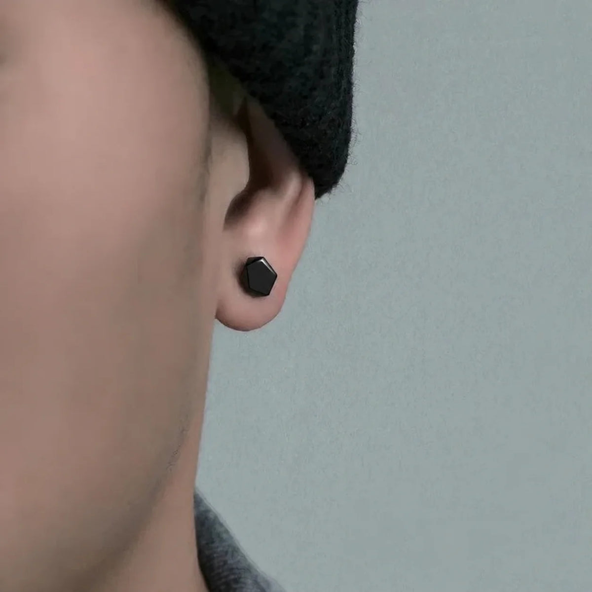 A close-up of a person's ear wearing a small matte black hexagonal stud earring, with part of a dark knit beanie and gray shirt visible against a soft light gray background.