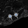 A pair of silver-toned starburst stud earrings with round light blue gemstones, displayed on a textured black rock surface.