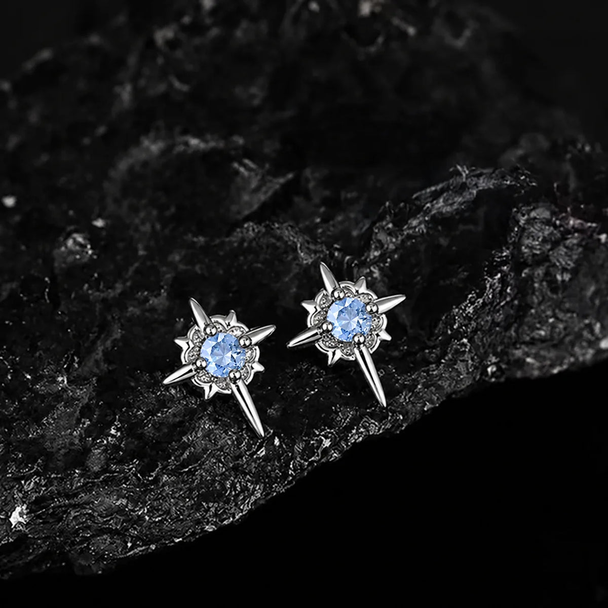A pair of silver-toned starburst stud earrings with round light blue gemstones, displayed on a textured black rock surface.