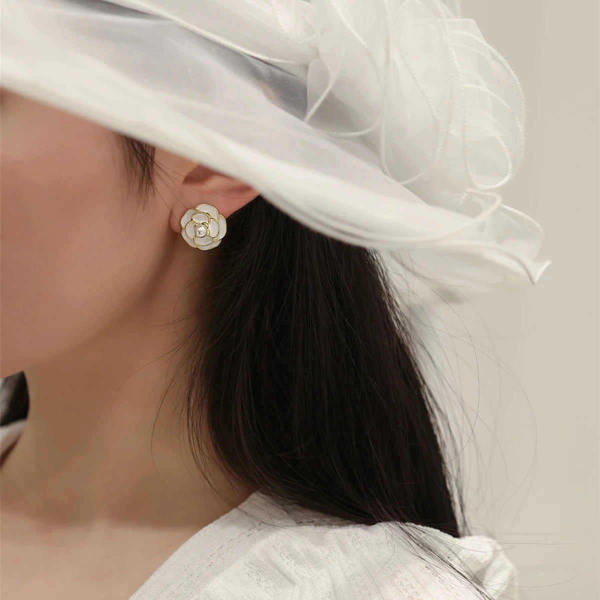 White Camellia enamel clip-on earrings worn on ear, side view with white hat and dark hair