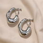 Silver chunky curved hoop clip-on earrings displayed on beige fabric showing polished metal finish and secure closure mechanism