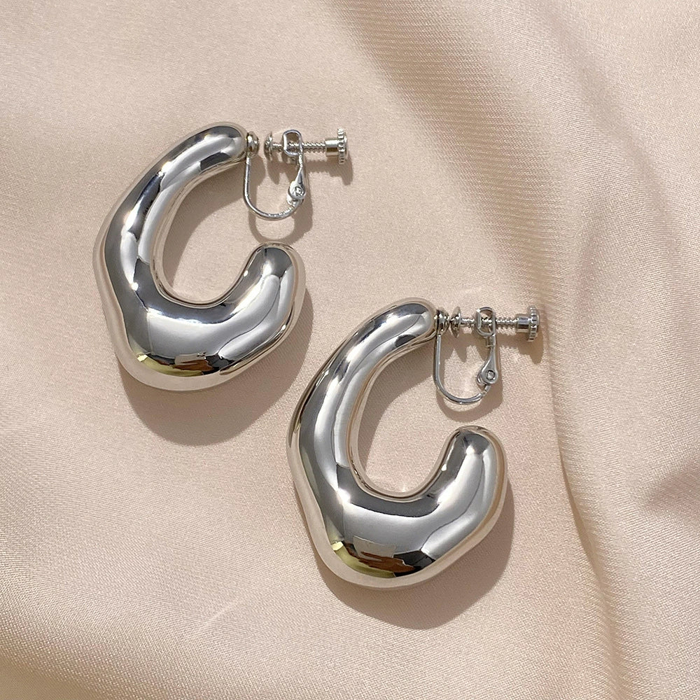 Silver chunky curved hoop clip-on earrings displayed on beige fabric showing polished metal finish and secure closure mechanism