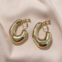 Chunky curved gold hoop clip-on earrings displayed on white fabric, showing polished metallic finish and elegant curved design from front angle