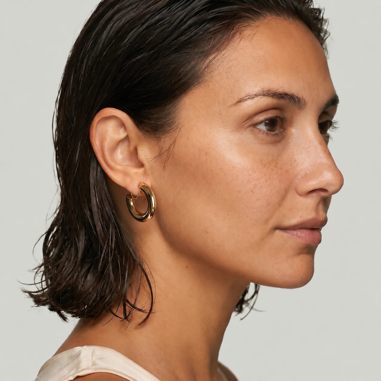 A close-up side profile of a woman wearing a pair of small gold hoop earrings against a light gray background.