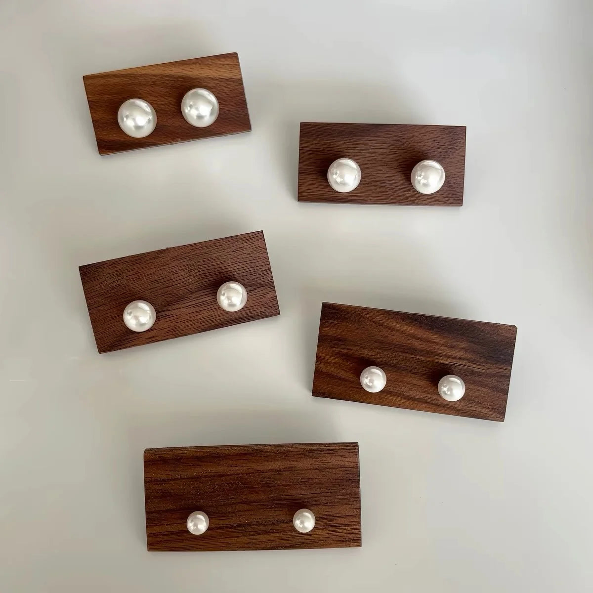 Five pairs of double stud earrings with white pearls set in dark brown wooden rectangular bezels, arranged on a light off-white background.