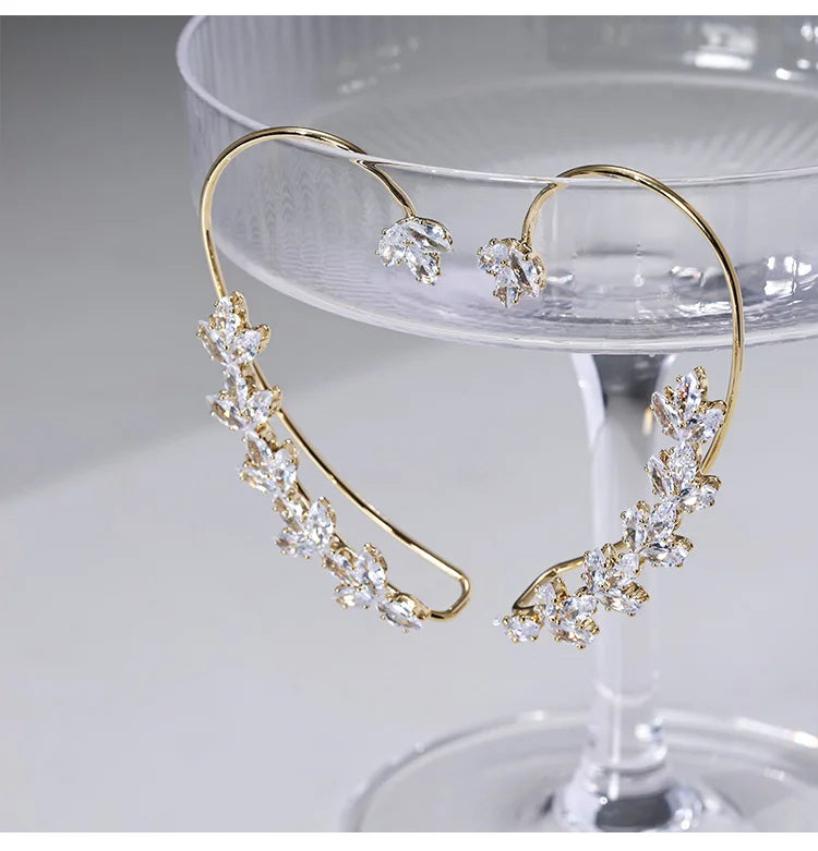 Crystal Leaf Cuff Earrings in Gold displayed on a champagne flute, showcasing the delicate cubic zirconia leaf design and curved gold wire structure from a front angle view.
