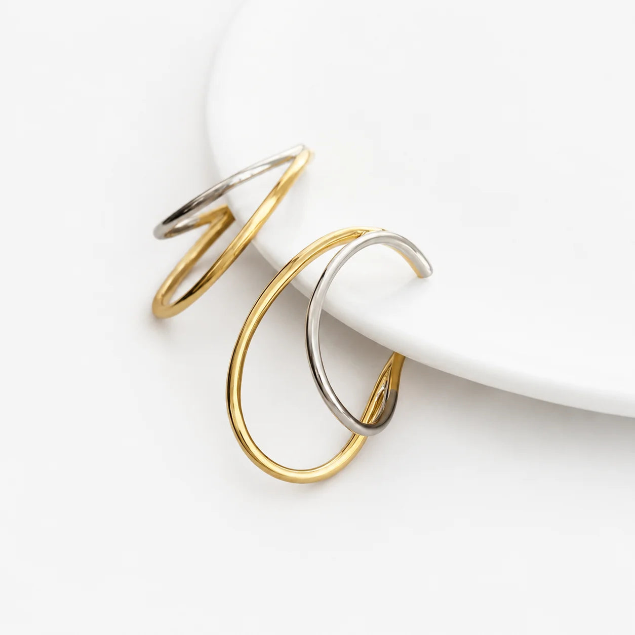 Two minimalist open hoop ear cuffs in polished yellow gold and silver, displayed on a clean white surface.