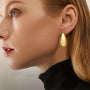 Vue de profil d'une femme portant des boucles d'oreilles pendantes minimalistes en or, de forme arrondie et épaisses, réalisées en alliage de cuivre doré avec une fermeture à charnière polie. Ces créoles apportent une touche chic à toute tenue moderne.