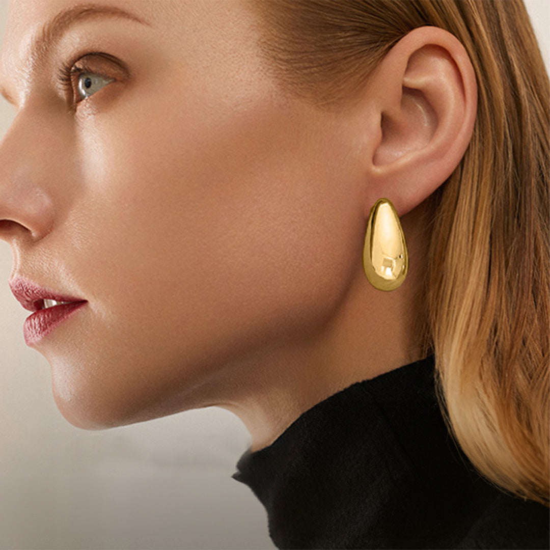 Profile view of a woman wearing minimalist gold chunky drop clip-on earrings, fashioned in a bean shape from a gold tone copper alloy with a polished hinge-back closure. These hoop-style earrings are a chic statement for any modern ensemble.