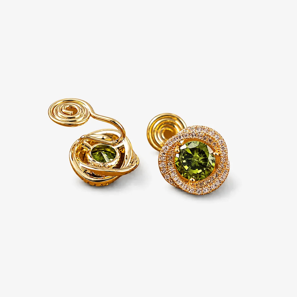 A pair of gold earrings featuring round green gemstones, one with a spiral twist design and the other with a halo of small clear stones, set against a light gray background.
