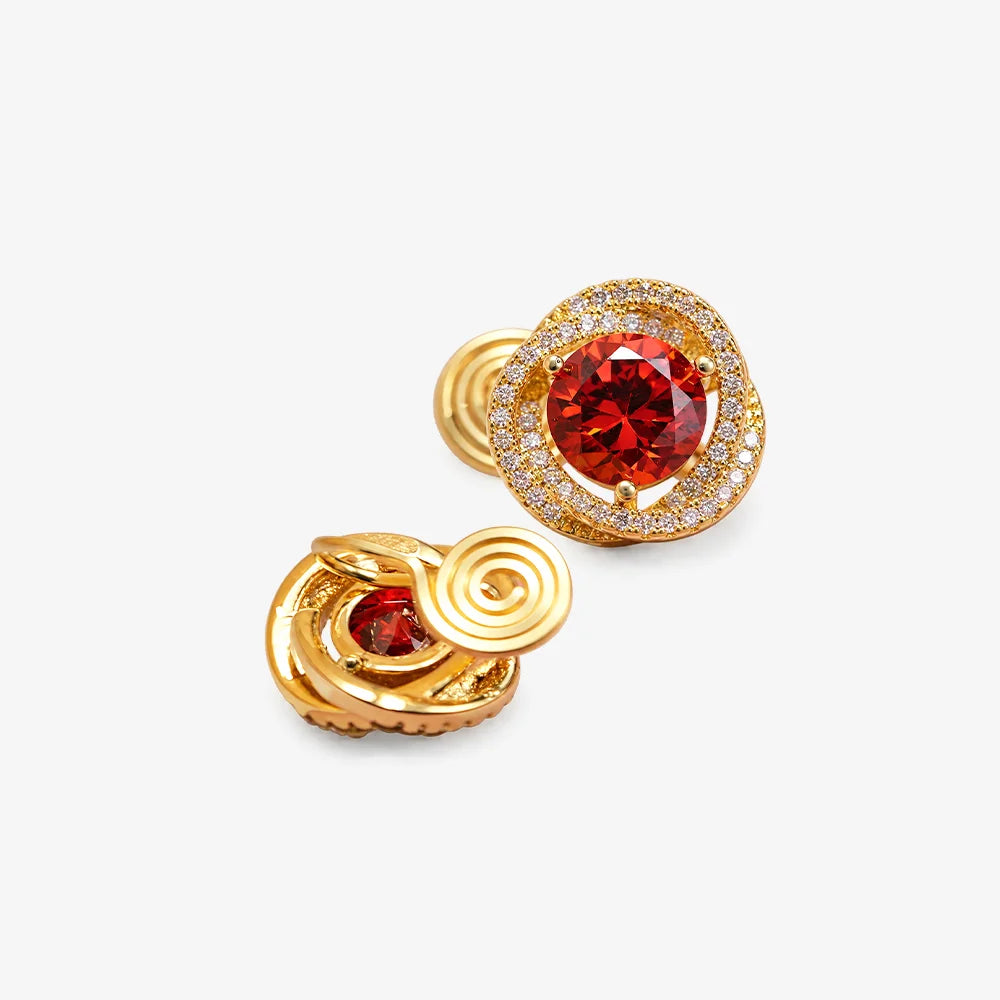 A pair of gold-plated stud earrings featuring a large round red gemstone in the center, surrounded by a halo of small clear stones, with an additional smaller red gemstone set in a swirling gold bezel on the reverse side, shown on a light gray background.