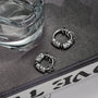 Silver Embrace clip-on hoop earrings displayed on dark surface next to water glass, showing detailed ornate design