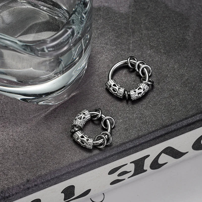 Silver Embrace clip-on hoop earrings displayed on dark surface next to water glass, showing detailed ornate design