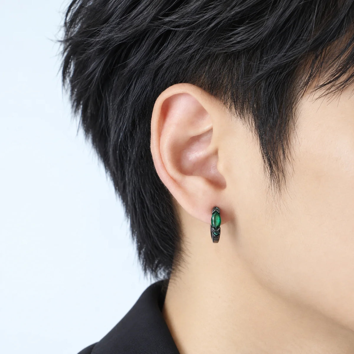 A close-up of a person's ear wearing a small black hoop earring with a green oval gemstone and textured detailing.