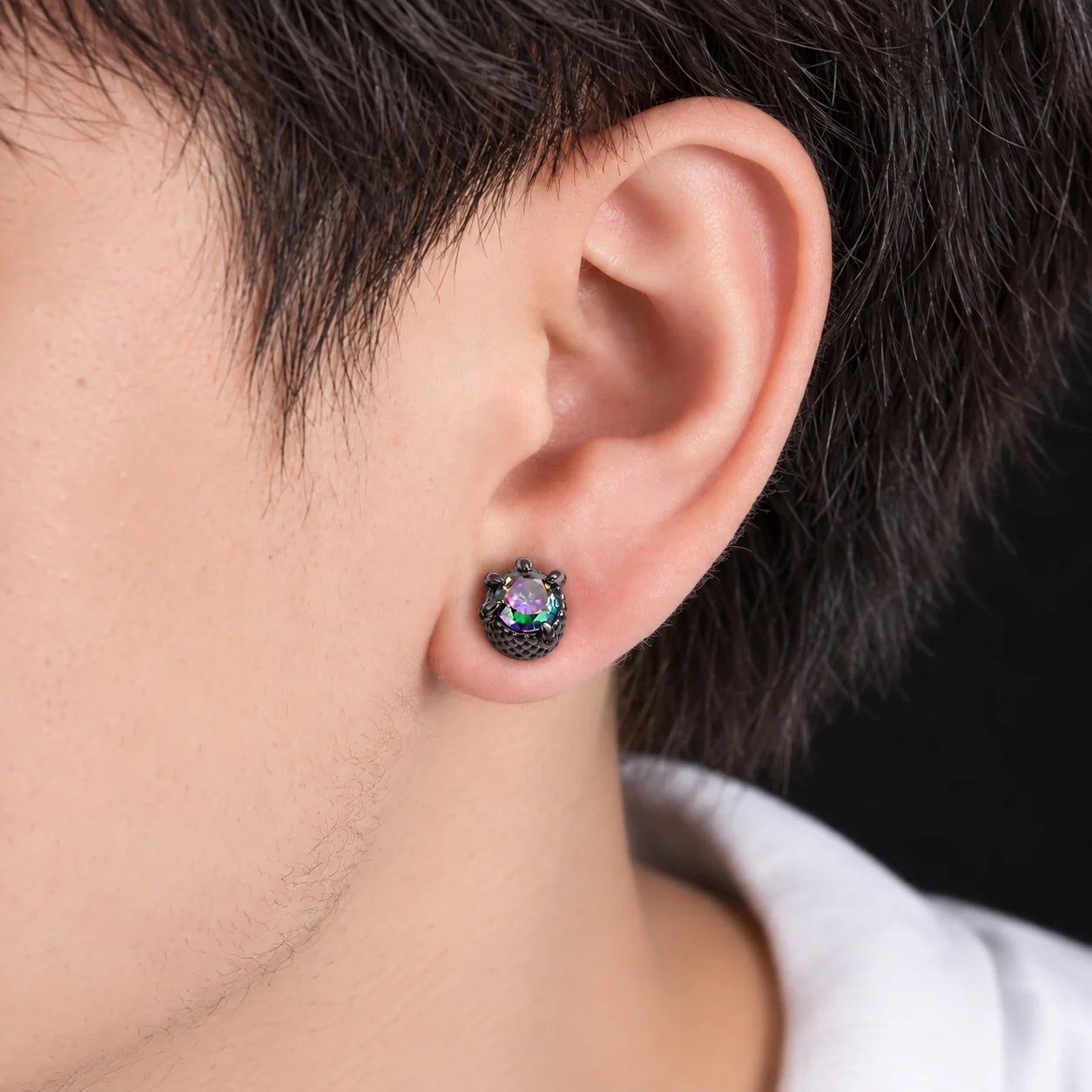 A close-up of a person's ear wearing a small stud earring with a dark textured setting and a faceted iridescent stone that shifts between green, purple, and blue.