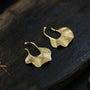 Gold ginkgo leaf ear cuff earrings displayed on dark wood surface, showing curved hook design and textured finish details.