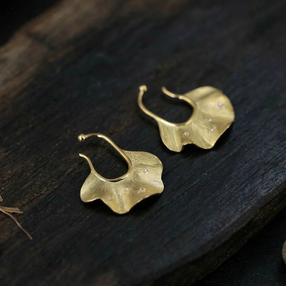 Gold ginkgo leaf ear cuff earrings displayed on dark wood surface, showing curved hook design and textured finish details.