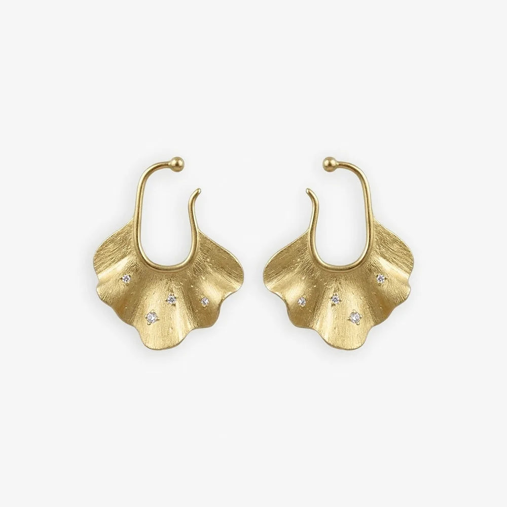 Boucles d'oreilles manchette en forme de feuille de ginkgo dorée, serties sur une surface blanche, ornées d'incrustations de zircon blanc étincelant qui captent la lumière, reflétant la beauté organique et le luxe des boucles d'oreilles manchette en forme de coquillage.