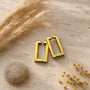 Gold rectangle clip-on hoop earrings displayed on neutral beige surface with decorative dried flowers and stone