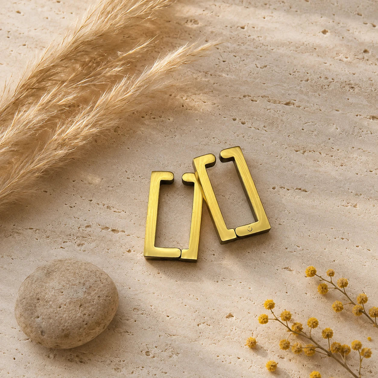 Gold rectangle clip-on hoop earrings displayed on neutral beige surface with decorative dried flowers and stone