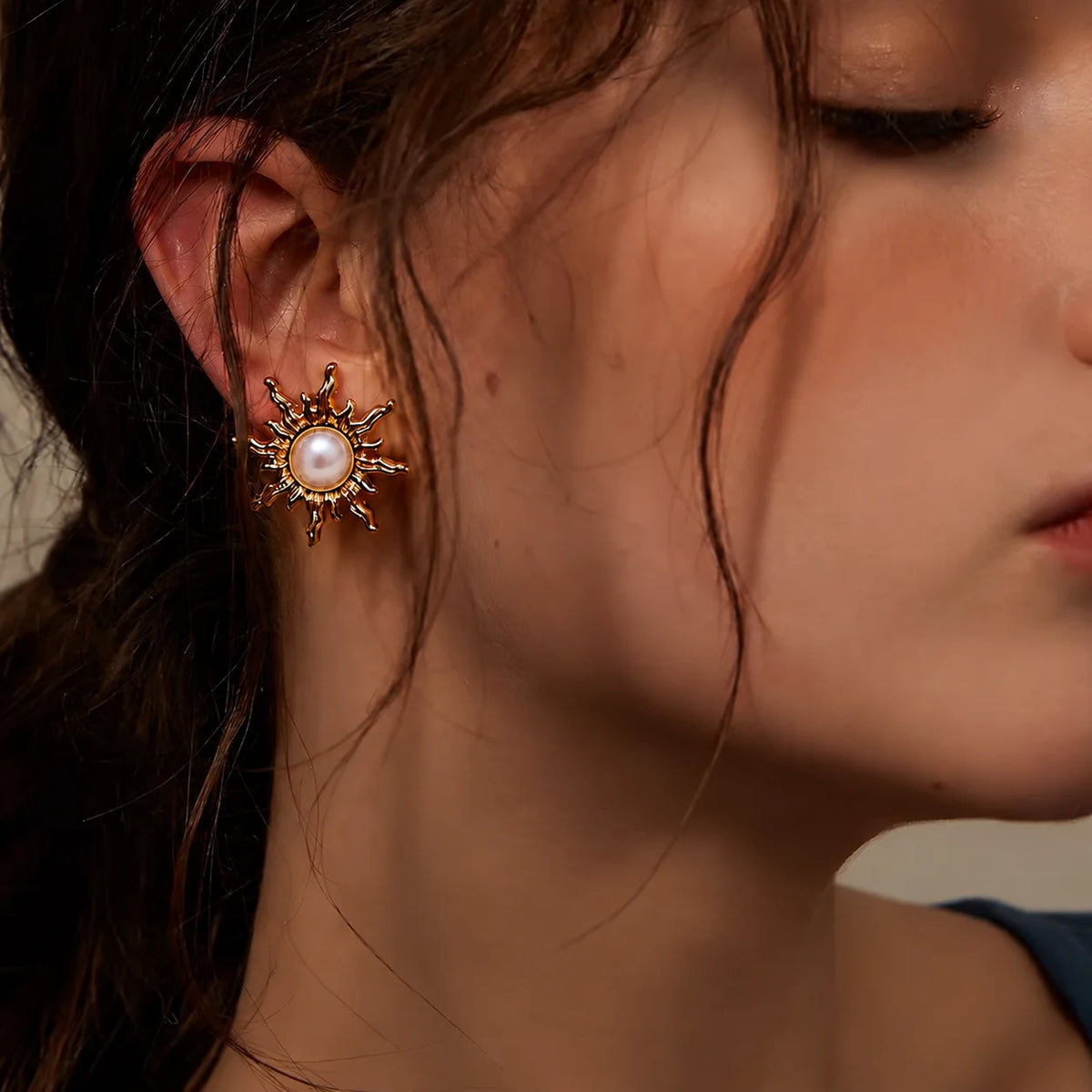 A close-up of a model's ear wearing a gold sunburst stud earring with a white pearl center and small clear stones.