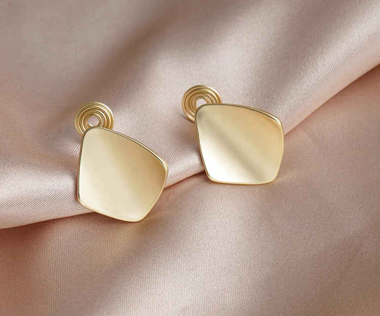 Gold square geometric clip on earrings displayed on beige fabric showing polished finish and gold-tone metal detail