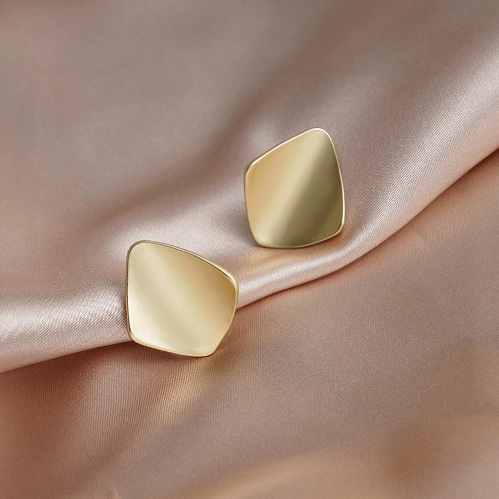 Gold square geometric clip on earrings displayed on beige fabric showing polished metallic finish and modern chunky design