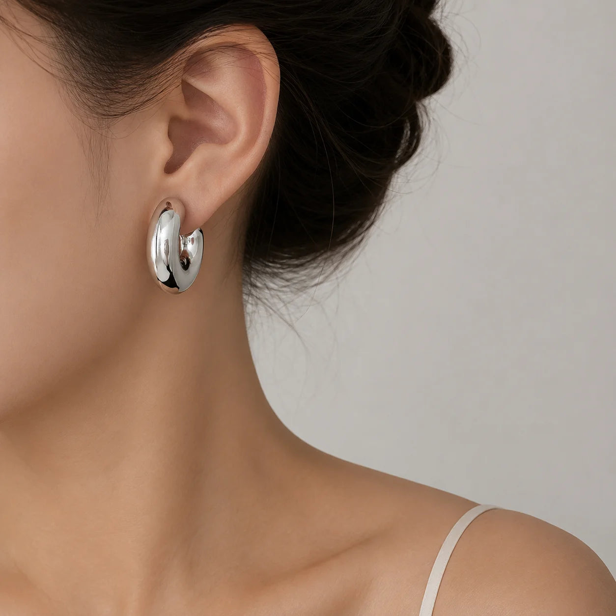 A close-up of a model's ear wearing a polished silver crescent-shaped hoop earring against a soft light gray background.