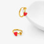 A pair of gold-toned hoop earrings, each featuring a small red heart-shaped enamel charm, displayed on a clean white background with a white ceramic dish prop.