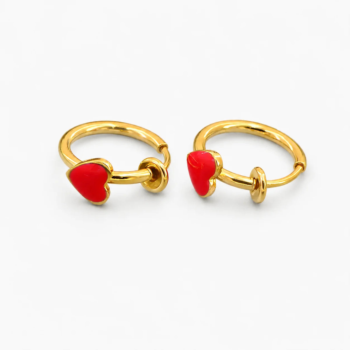 A pair of gold huggie hoop earrings, each adorned with a small red heart charm, displayed on a clean white background.