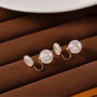 Iridescent pearl clip-on earrings displayed on brown fabric, showing lustrous white spheres with gold-toned clips from a side angle view
