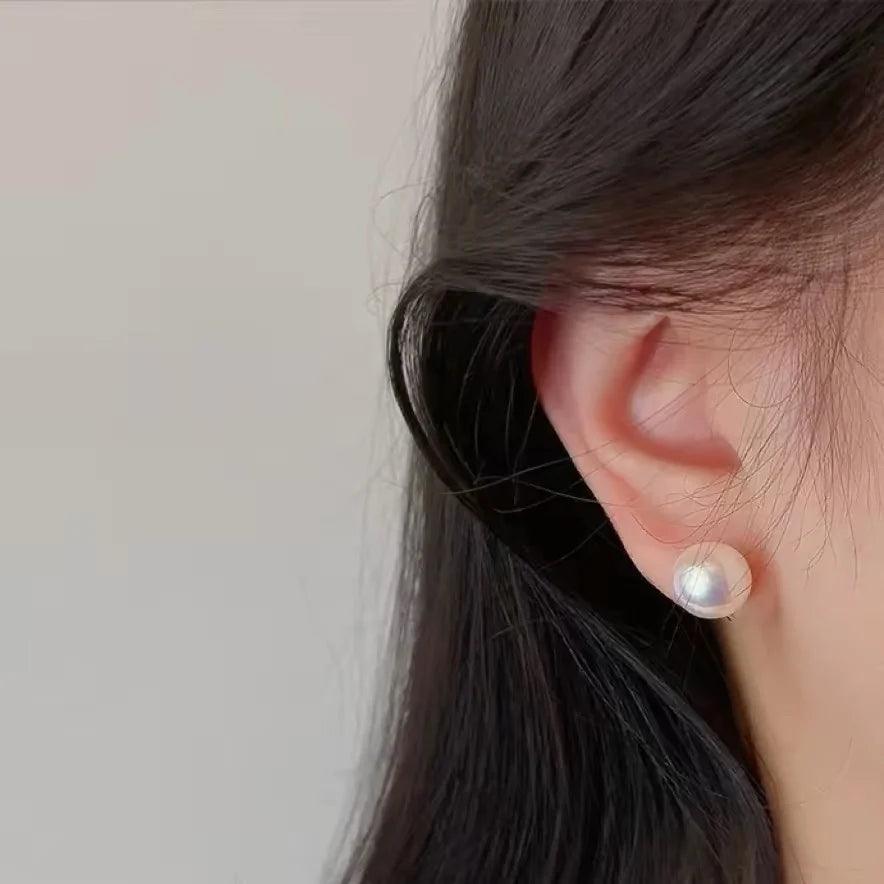 A close-up side view of a person's ear wearing a single round white pearl stud earring with a subtle gold setting, against a soft light gray background.