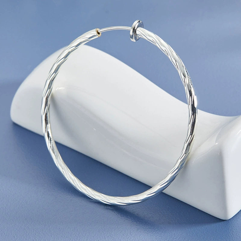 Large twisted textured silver hoop clip-on earrings displayed on white ceramic stand against blue background