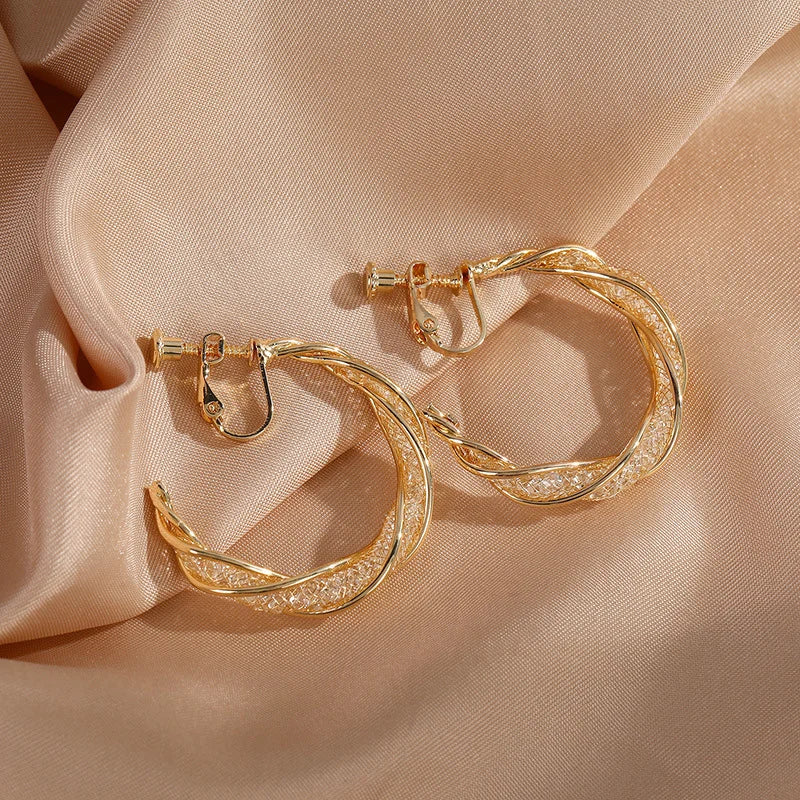 Gold glimmering lattice twist clip-on earrings displayed on beige fabric background, showcasing textured hoops from front angle