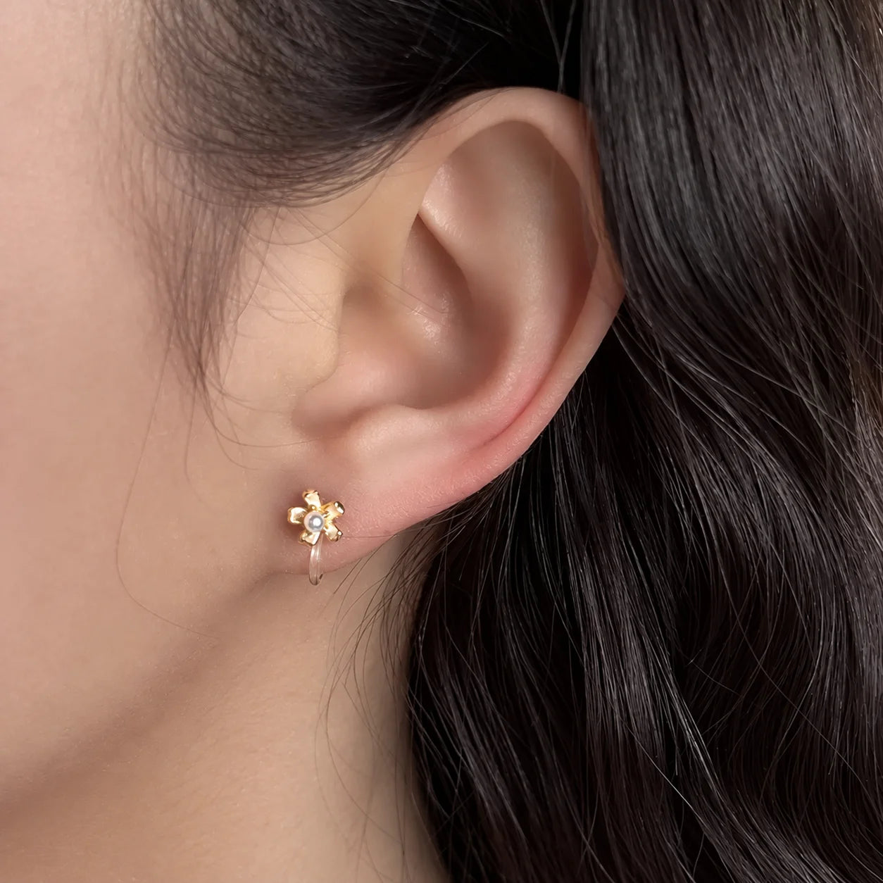 A close-up of a person's ear wearing a small gold flower-shaped stud earring with a tiny pearl in the center.