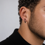 A close-up side profile of a man's ear wearing a simple black hoop earring, with curly dark hair and a black ribbed turtleneck against a light gray background.