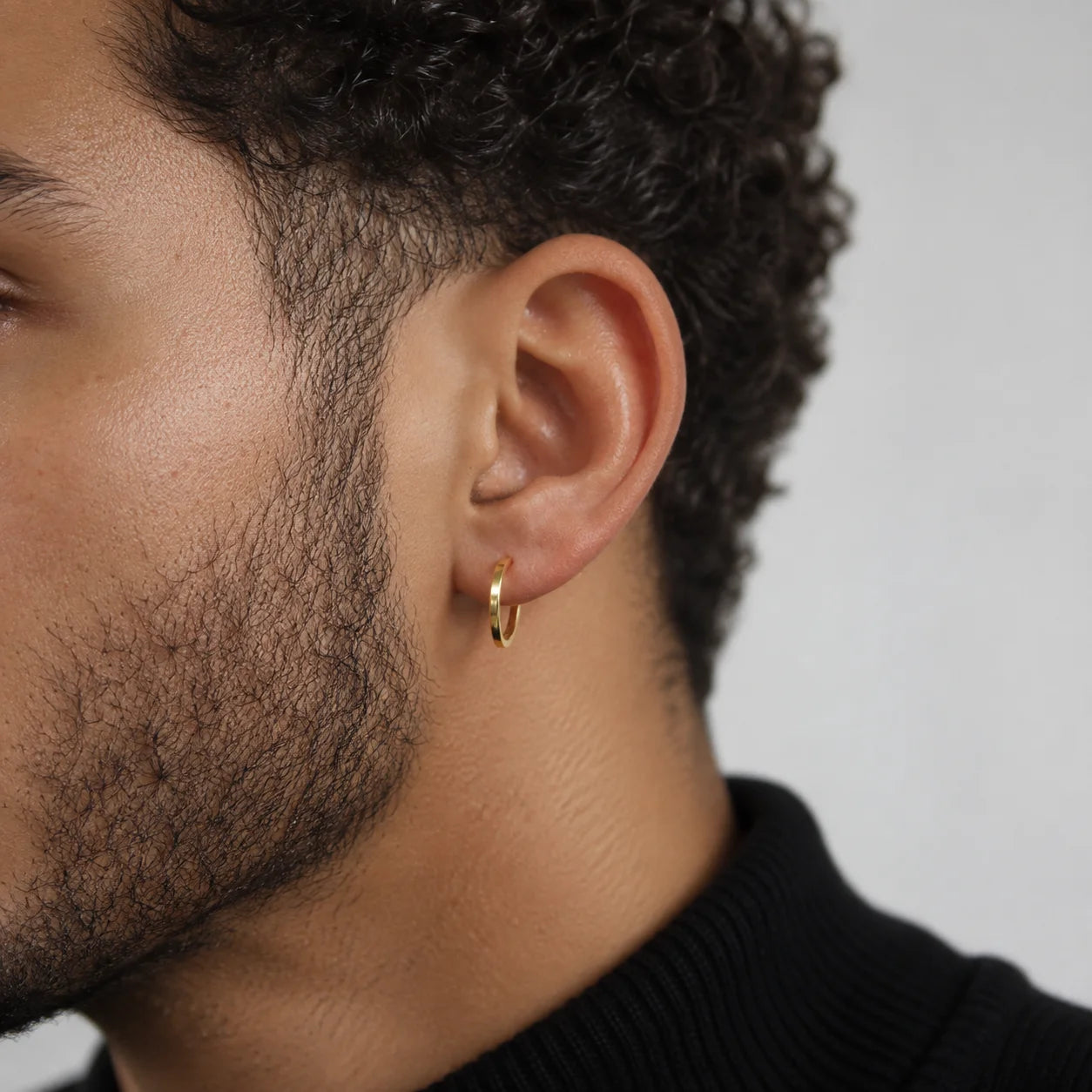 A close-up side profile of a man wearing a small gold hoop earring on his earlobe, against a plain light gray background.